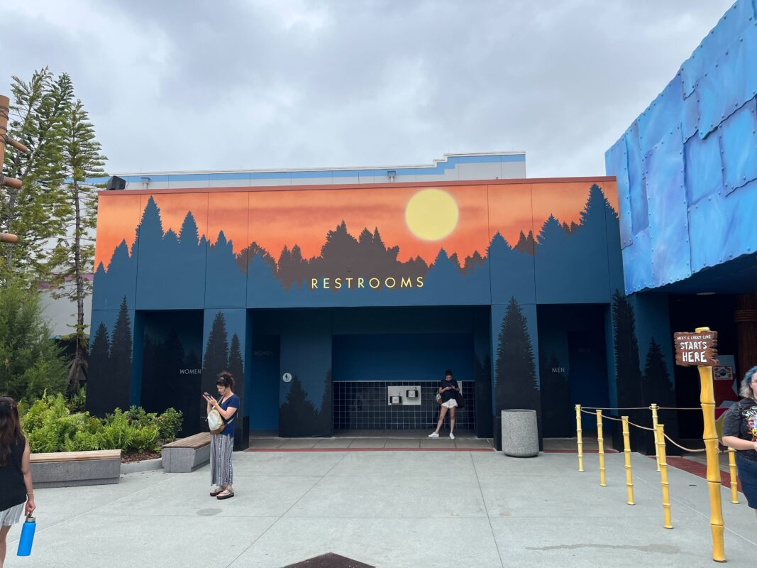 An outdoor restroom facility at Universal Studios Florida featuring an artistic forest mural alongside an E.T. mural and a "Restrooms" sign. Yellow barriers form a line on the right, and a few people are visible in front of the building.