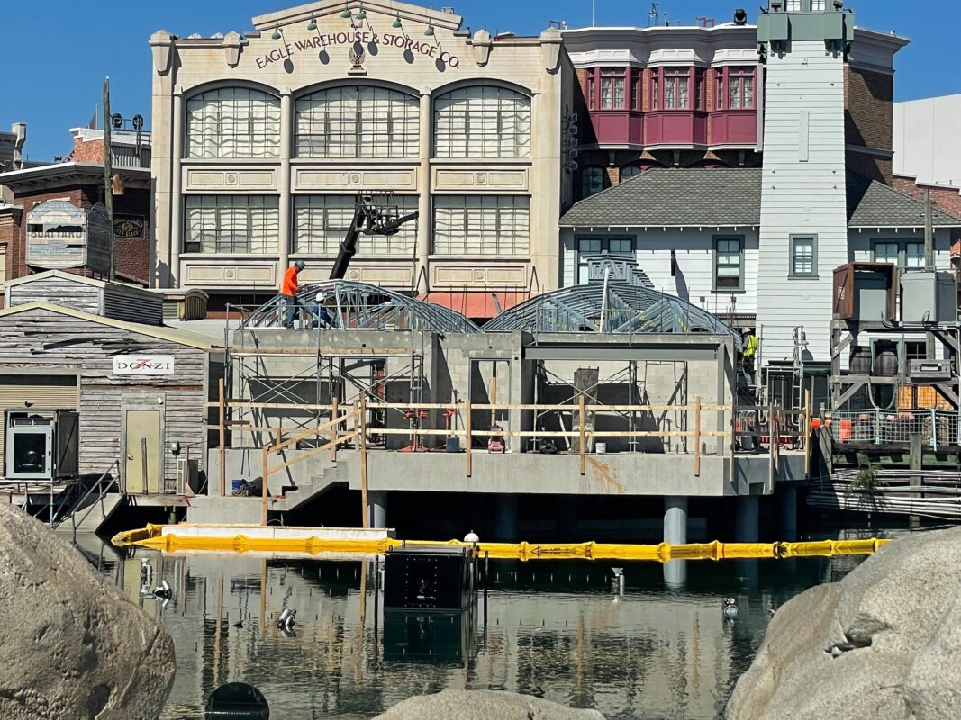 Construction workers building a metallic structure on a platform over a body of water, surrounded by buildings labeled Eagle Warehouse & Storage Co. and Docks. Yellow barriers are placed in the water.