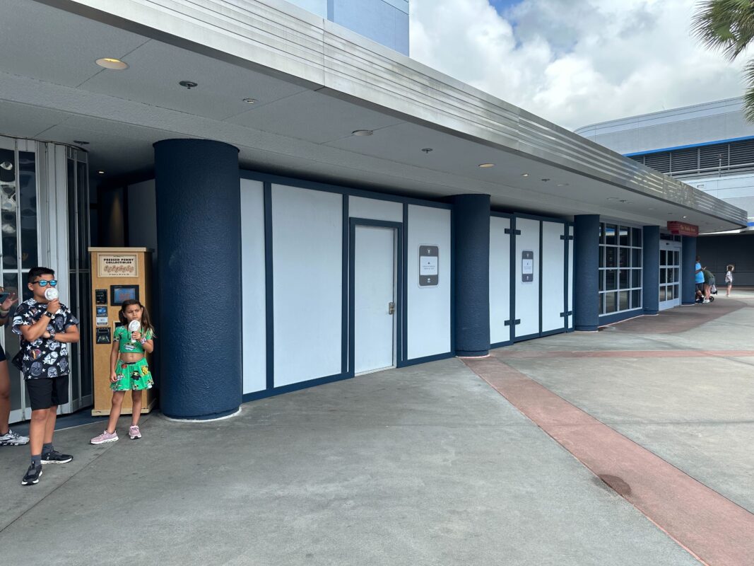 Two children stand near a blue and white building facade under a covered walkway. A closed door and construction barriers are visible. Cloudy skies are in the background.