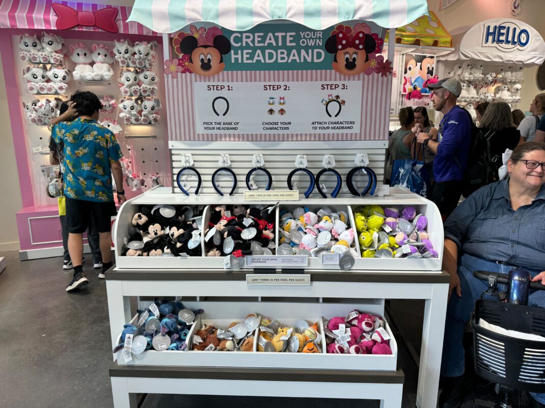 A shop display with multiple plush character headbands and accessories. A sign reads "Create Your Own Headband" with steps to pick a headband, choose characters, and attach characters to the headband.
