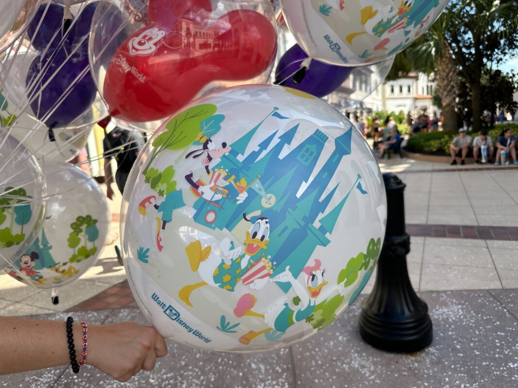 A person holding a bundle of colorful balloons featuring Disney characters including Donald Duck and Goofy, with people in the background at an outdoor setting.