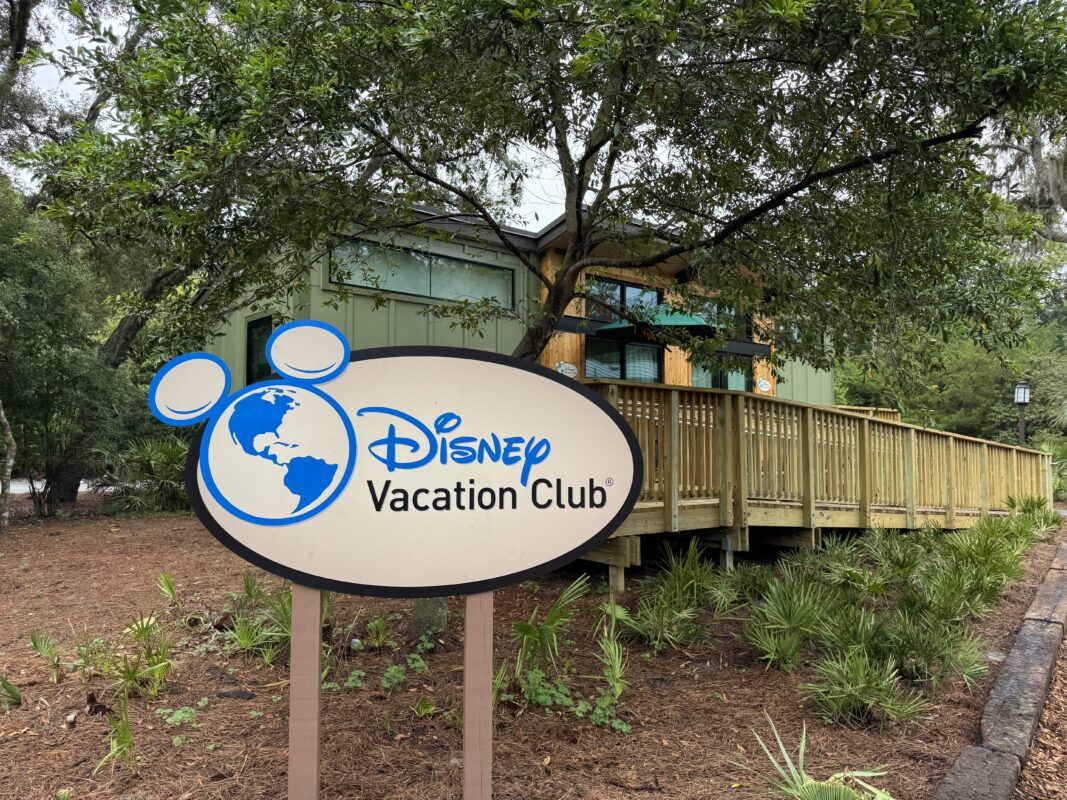 Sign that reads "Disney Vacation Club" in front of a green building surrounded by trees and shrubs.