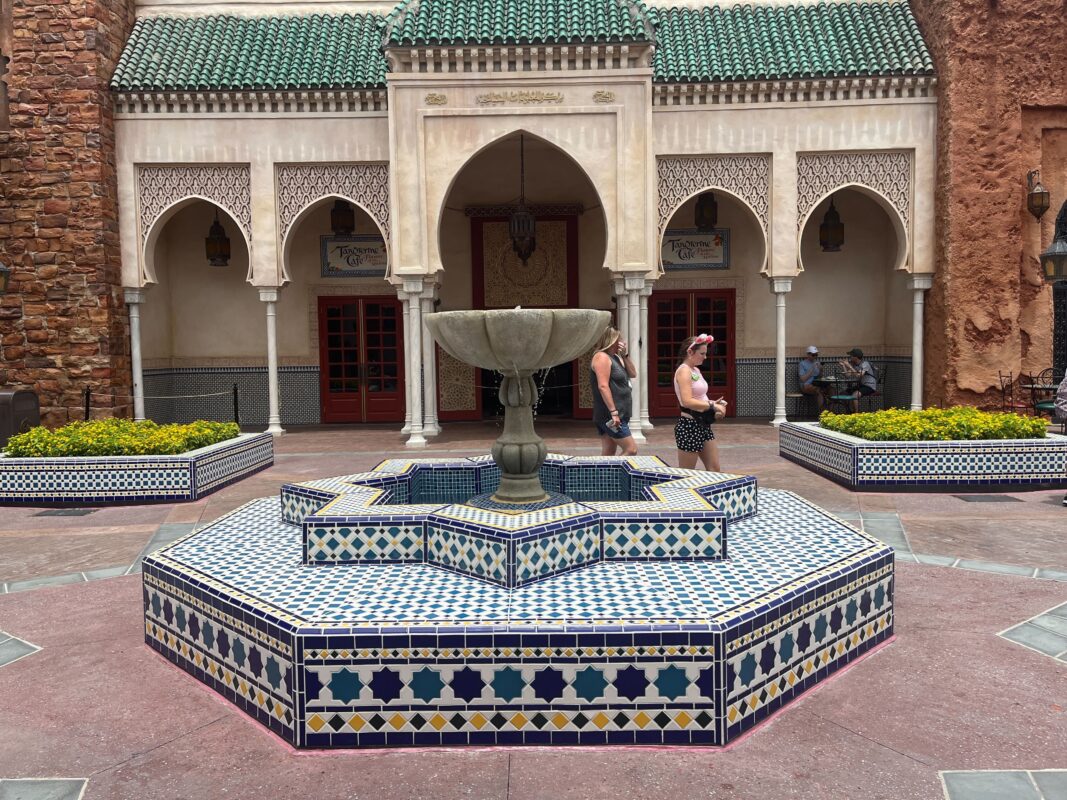 A tiled octagonal fountain with a central pillar stands in front of a building with archways and a green-tiled roof, reminiscent of the Morocco Pavilion. Two people walk nearby, and there are plants on either side of the fountain.