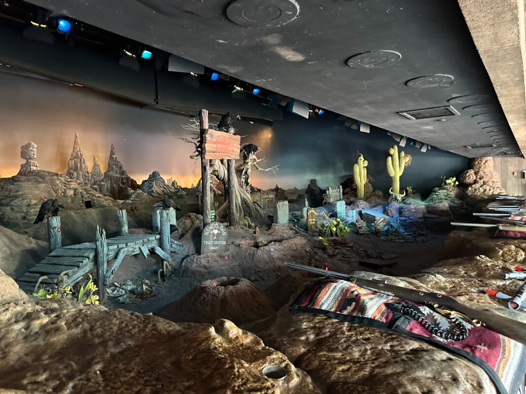 A detailed miniature diorama depicts an old Western desert scene with cacti, tombstones, a cross, and a wooden sign. The backdrop shows a twilight sky and rocky formations.
