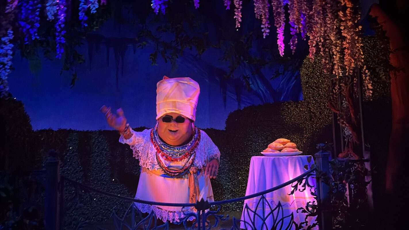 Mama Odie in a white dress and multiple necklaces stands next to a table with beignets, under purple flowering vines in a dimly lit, decorative setting.