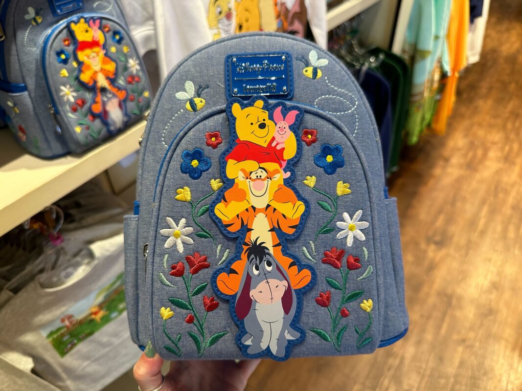 A blue backpack featuring embroidered characters from Winnie the Pooh stacked vertically: Pooh, Piglet, Tigger, and Eeyore, surrounded by colorful flowers.