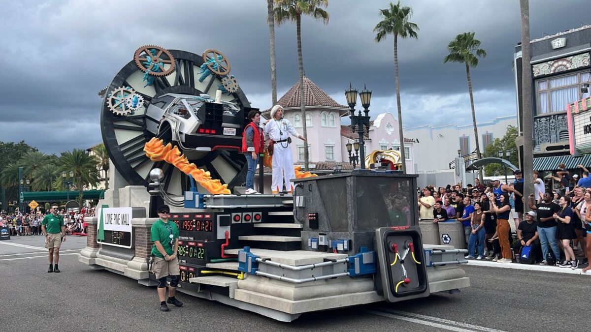 Parade float depicting a "Back to the Future" scene with a DeLorean car, fire effects, and characters in costume. The crowd excitedly watches the universal mega movie parade unfold against a cloudy backdrop.