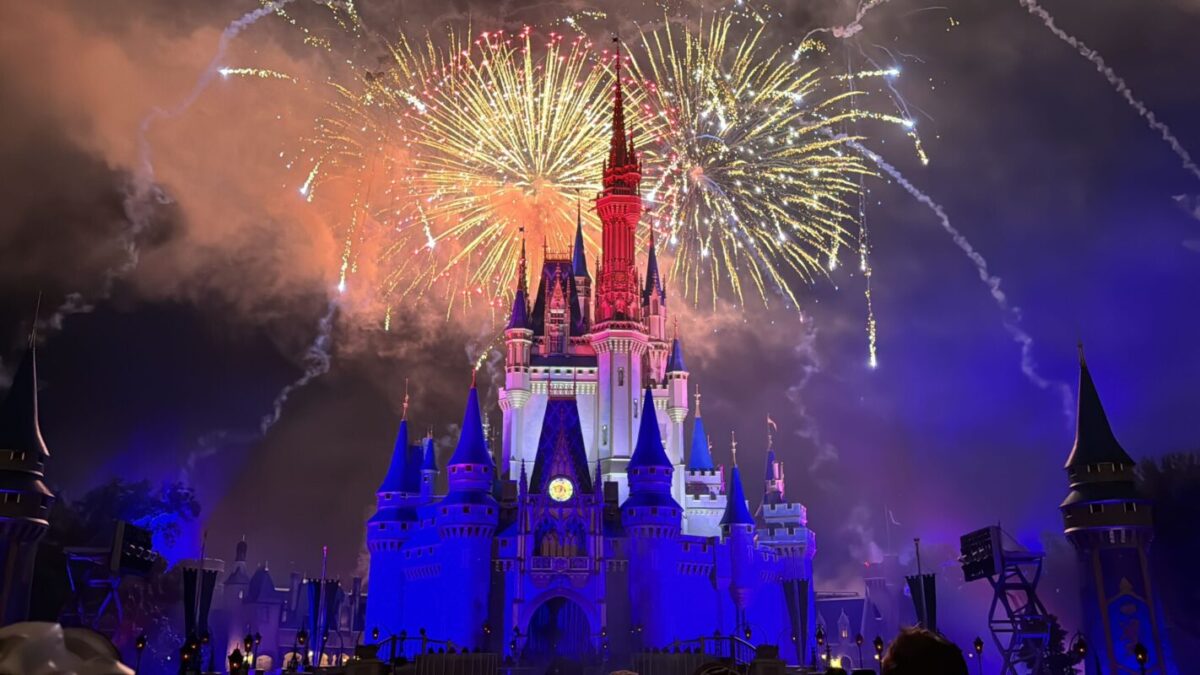 Fireworks illuminate the sky above a large, brightly lit castle at night, casting streaks of light and explosions in varying colors. Celebrating Fourth of July, the castle is surrounded by a crowd of enthusiastic people.