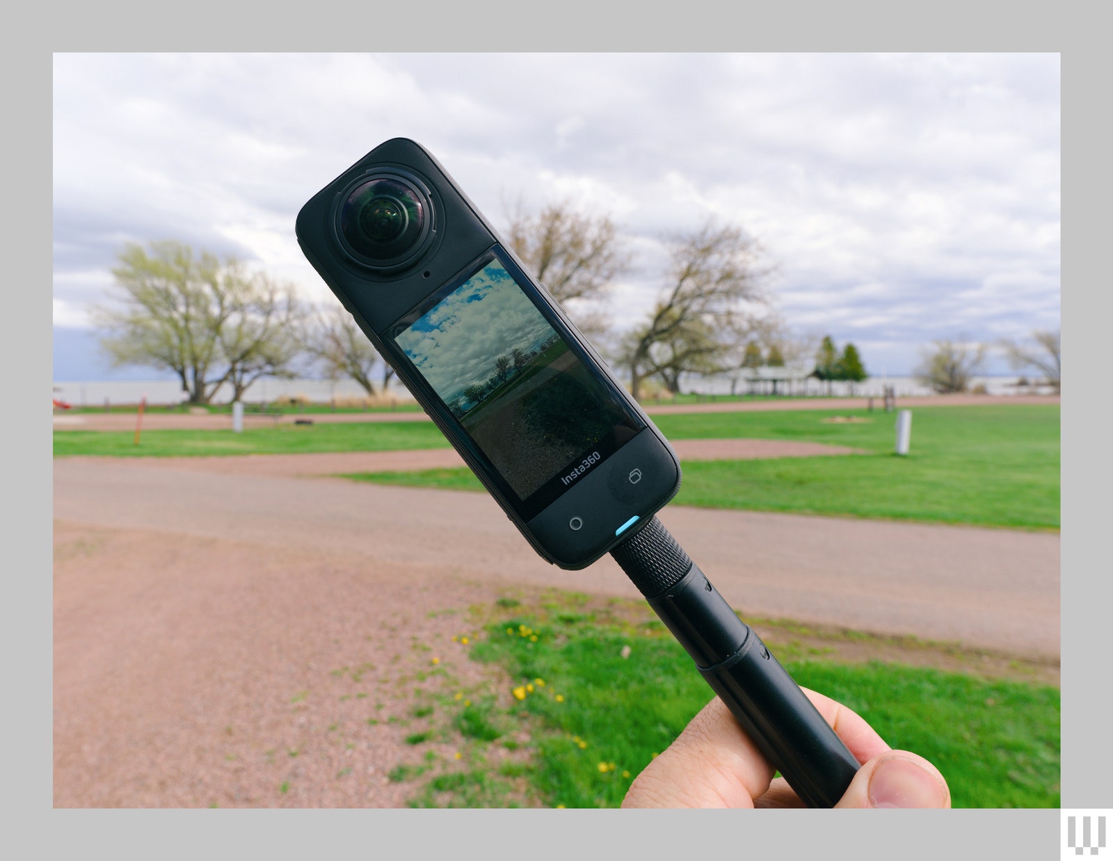 Black rectangular digital camera at an angle attached to a rod with the screen capturing the clouds and trees in the...