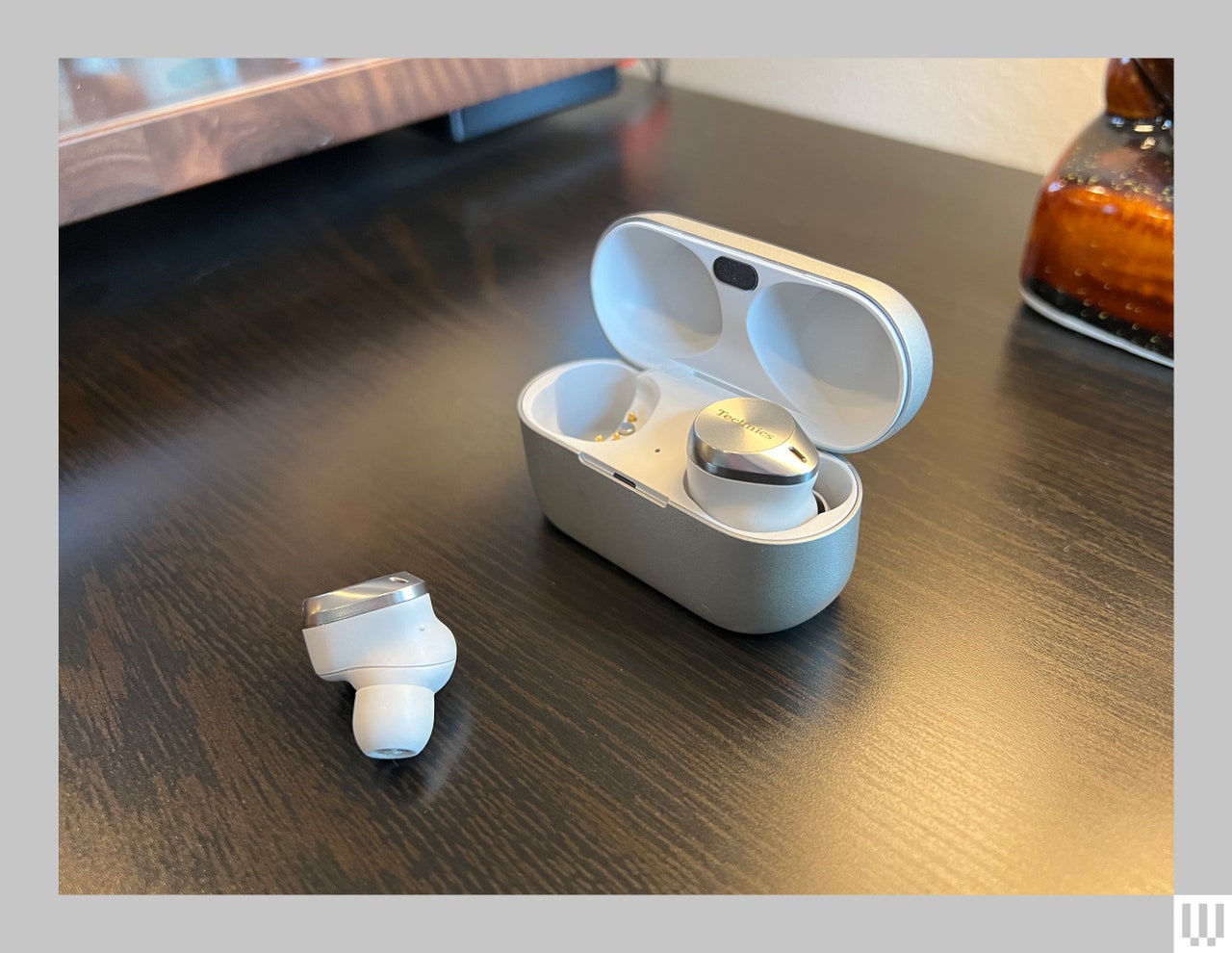 Oval-shaped case with white interior holding an earbud with the other earbud on the table beside the case 