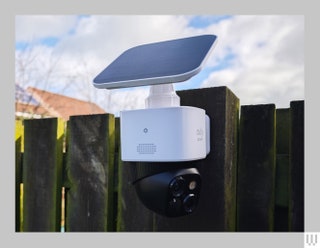 Two part outdoor security camera system attached to a fence. The top is a white box with extended solar panel. The...