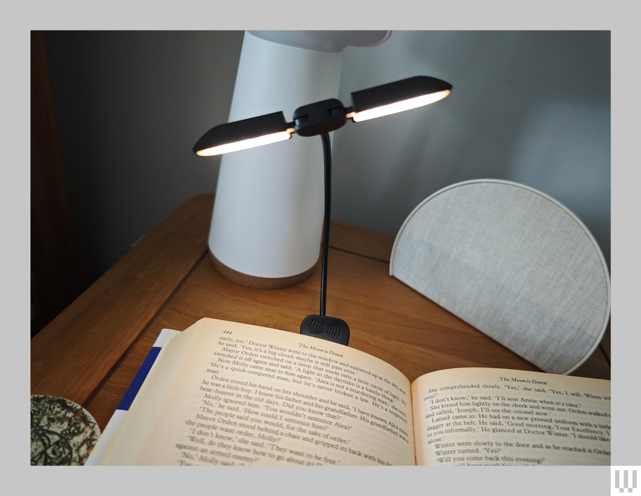 A book opened on a wooden nightstand with a thin two-sided light clipped to the book