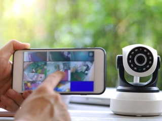 Hand holding a mobile phone with the screen showing live video feeds from security cameras within a home. A white...