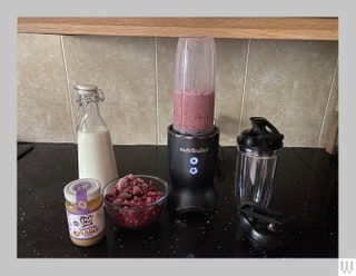 Small blender with black base sitting beside ingredients and an extra container all on a black kitchen counter