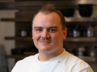 Headshot of Chef Garrett Rochowiak a person with short brown hair wearing a white chef jacket with utensils and cookware...