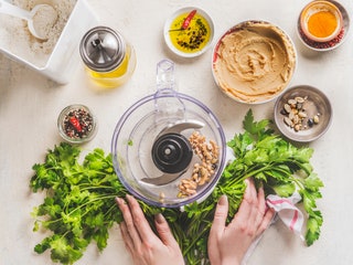 Overhead view of two hands holding leafy greens near a food processor with ingredients surrounding such as nuts oil and...