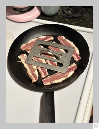 Lodge 12Inch Carbon Steel Skillet on a white stove with strips of bacon inside while a metal plate with slots and a...