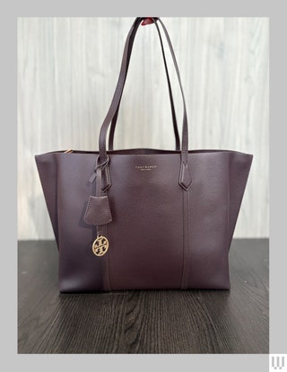 Tory Burch Perry TripleCompartment Tote Bag in brown with two skinny handles tugged upwards and a gold charm dangling...