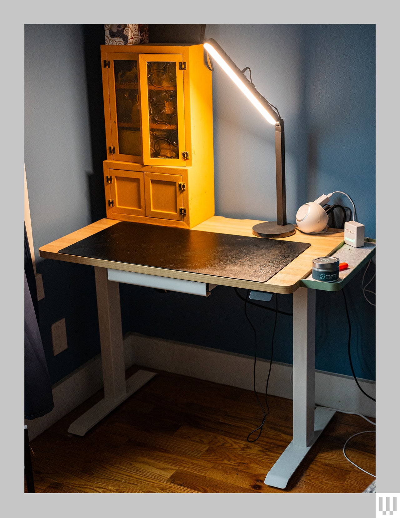 Branch Duo Standing Desk with light brown top and white legs that has a small cabinet, black cutting mat, and lamp on the top