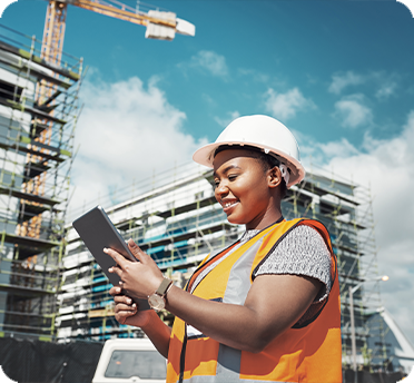 Woman in an orange safety vest and hard hat at a construction site looking at a clipboard