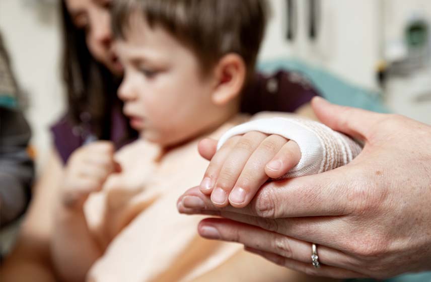 Toddler getting his hand/wrist bandaged with a cast while sitting on a woman's lap.