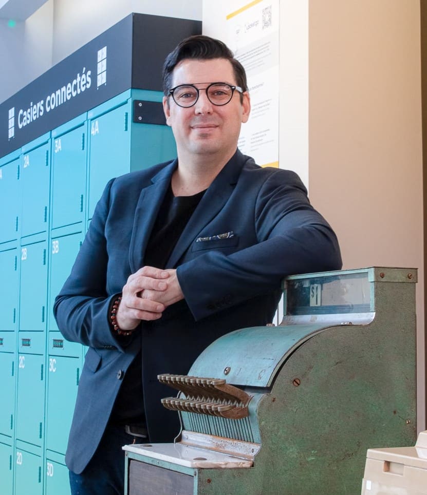 Carl Boutet positioned between a classic cash register and modern collection lockers for online ordering