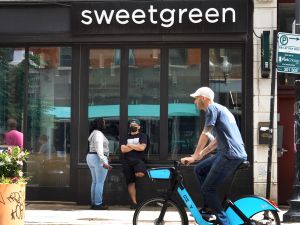 Customers stand in front of a Sweetgreen chain storefront while another person rides a bike in the opposite direction.