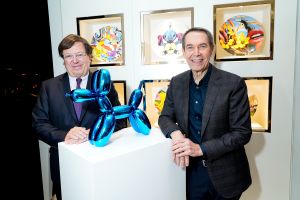 Jeff Koons poses in gallery next to blue balloon dog atop white mantle.