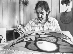 Black and white image of Norval Morrisseau sitting at a desk, painting canvas laid out in front of him
