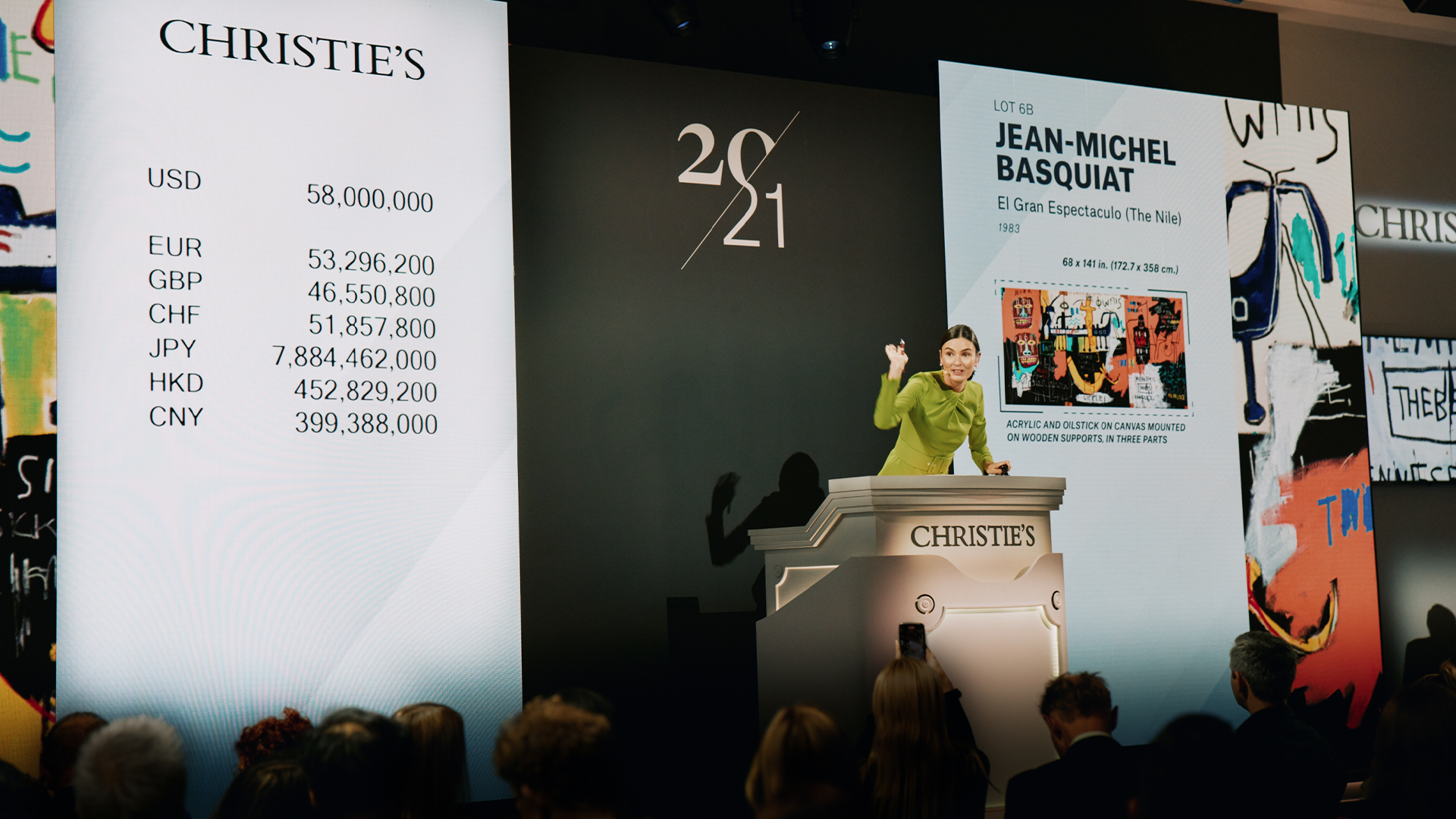 A woman in a green dress stands at a dais leading an auction with a large electronic display behind her.