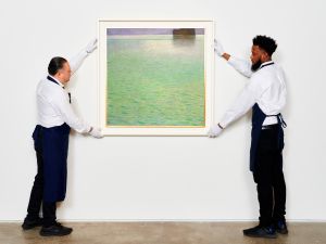 Two men with aprons and gloves hold up a framed painting of an ocean.