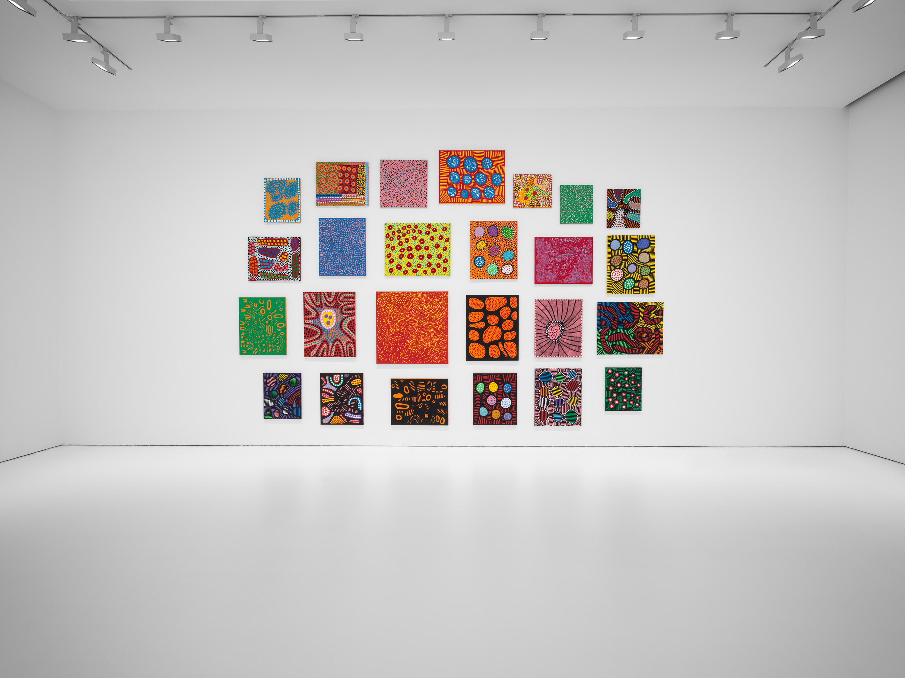 A stark art gallery space hosts a wall of small colorful paintings featuring Yayoi Kusama's signature dots.