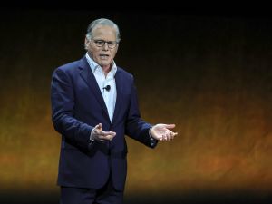 David Zaslav stands on stage wearing a suit with his hands out.
