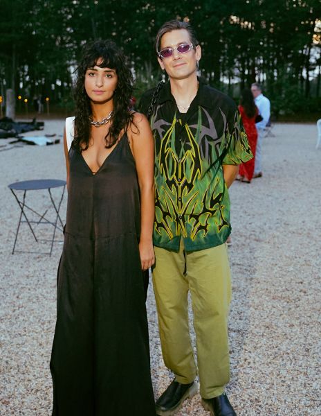 A woman in a black dress and a man in sunglasses, a green flame decal shirt, and green pants.