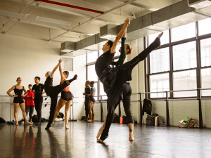 Ballet dancers in a high arabesque rehearse in a busy studio