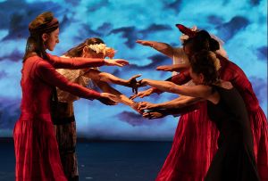 Dancers in red costumes reach their hands toward each other on a stage lit in blue