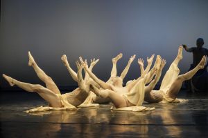 A group of painted dancers lies in a circle on a stage with their legs and arms in the air