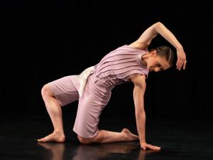 A dancer in a knee-length costume stretches over her own body on a stage