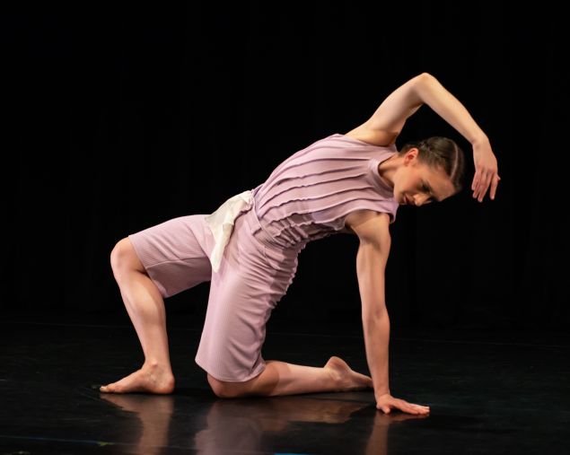 A dancer in a knee-length costume stretches over her own body on a stage
