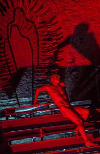 A nude dancer lit in red lounges on a stage