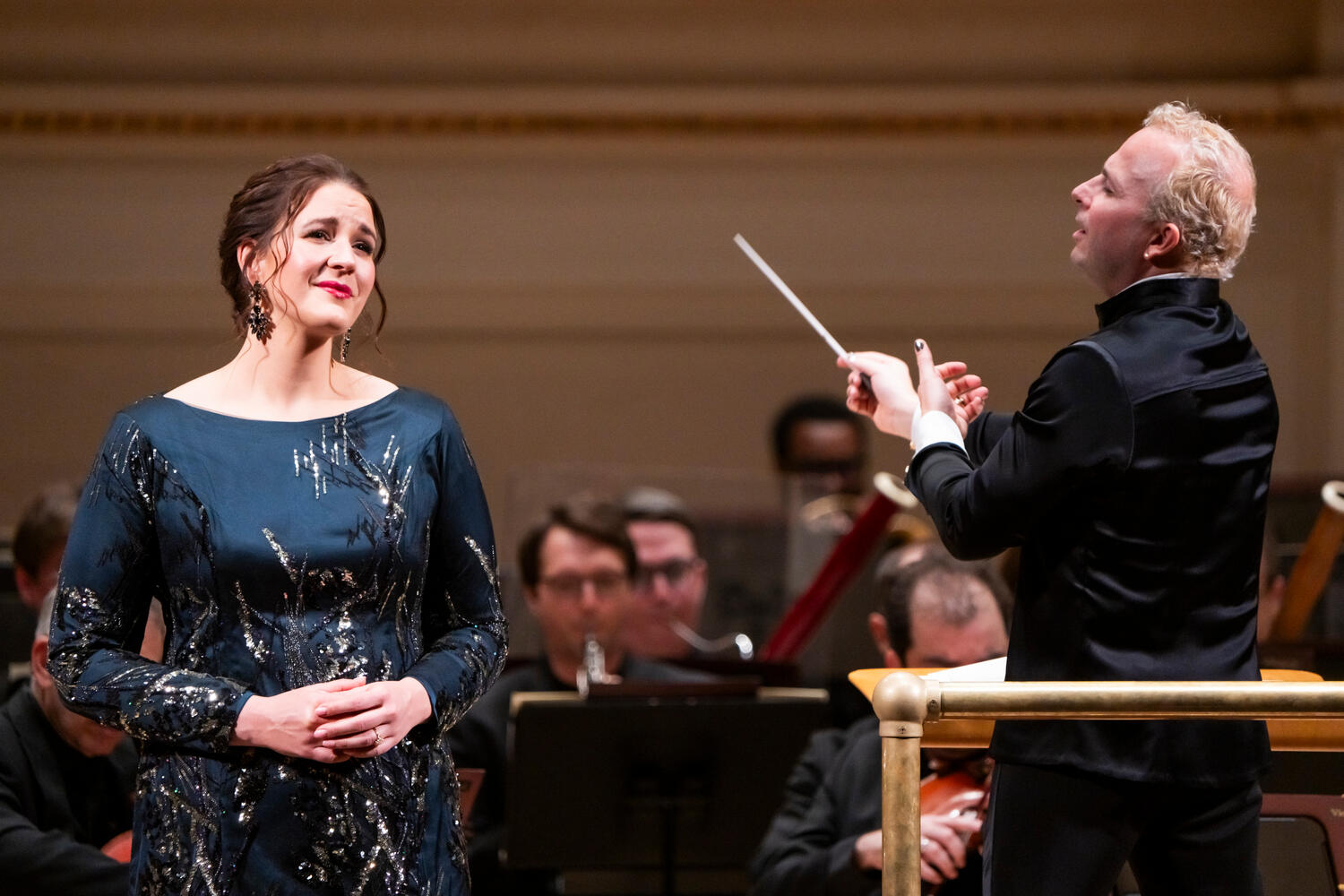 A woman in a blue dress sings while a conductor conducts