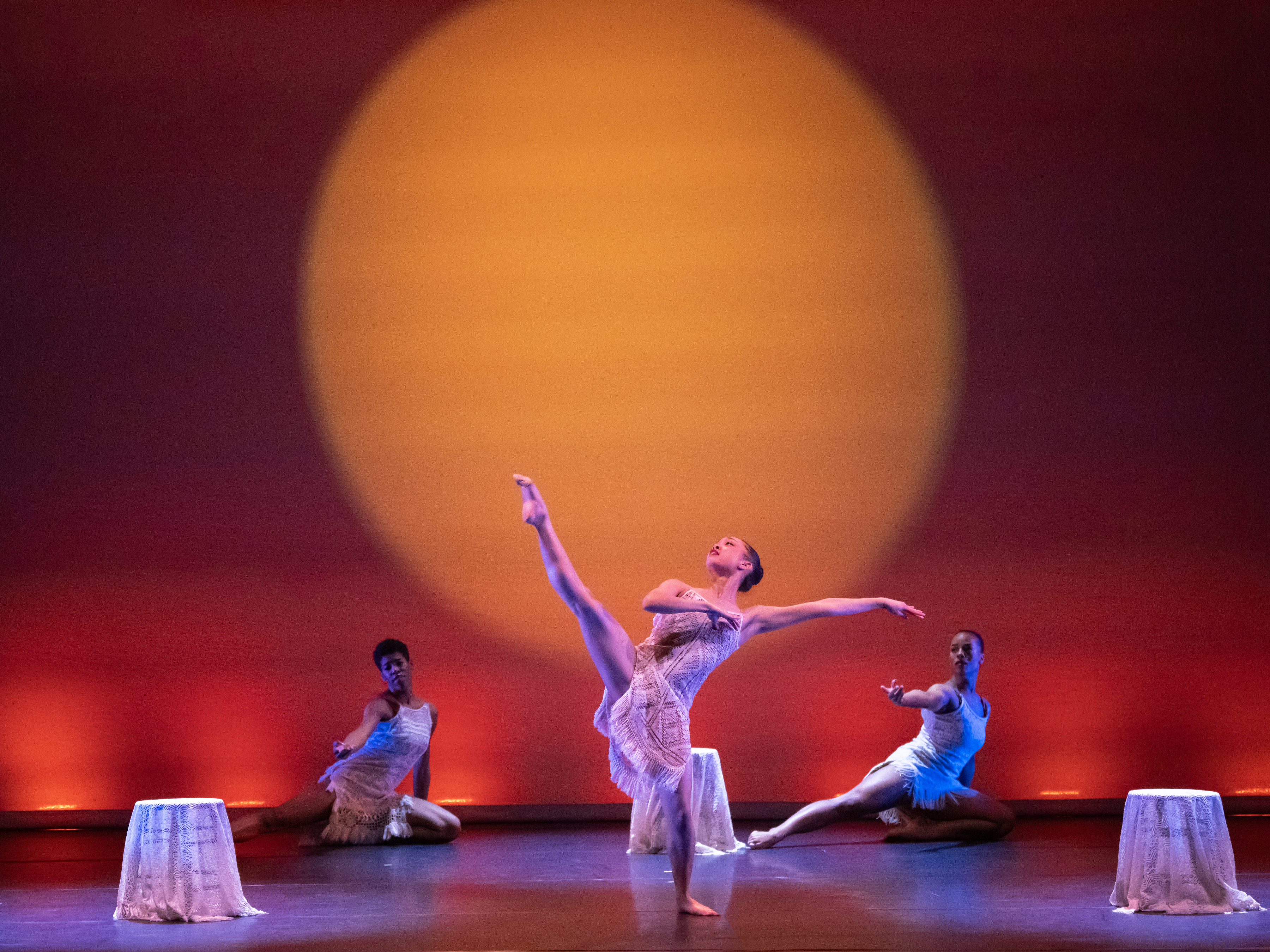A dancer kicks while two other dancers crouch on a stage with a backdrop that looks like a setting sun