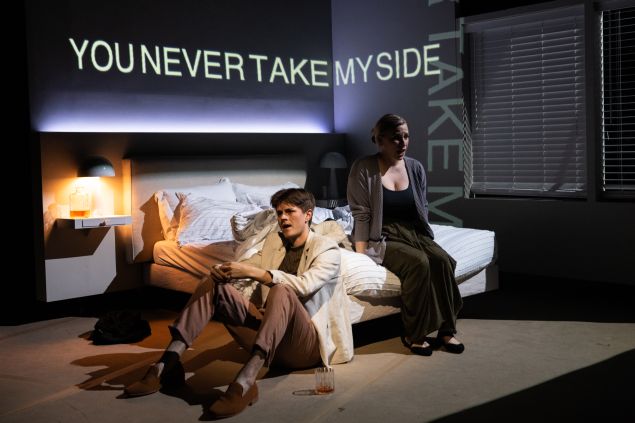 Two performers sit, one on the floor and one on a bed, on a stage set to look like a bedroom