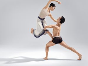 A male dancer in black dance briefs lifts a female dancer in a white dress with a black belt