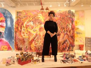 An artist stands among paints and brushes in front of a large and colorful painting