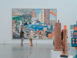 Two people stand in front of a large painting of a blue car in a bright gallery space