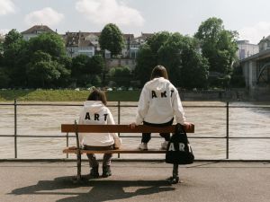 Two girls sit on park bench wearing hoodies that read "ART" on the back