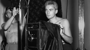 A shirtless man stands behind a large old fashioned camera while another shirtless man looks on