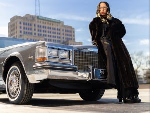 A woman wearing a long coat leans on the hood of an old car with her arms crossed; she's staring at the viewer