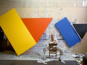 A man sits in a chair in front of an artwork made of colorful shapes